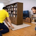 黄色のシャツを着たスタッフが棚を動かすのに布を引っ張っている。隣の女性が驚いた表情でその様子を見守っている