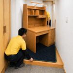 玄関先の狭い廊下で、スタッフ2名が大型の木製学習机を養生マットの上に載せて慎重に搬入している様子