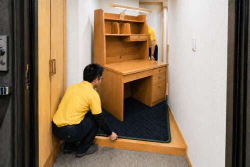 玄関先の狭い廊下で、スタッフ2名が大型の木製学習机を養生マットの上に載せて慎重に搬入している様子